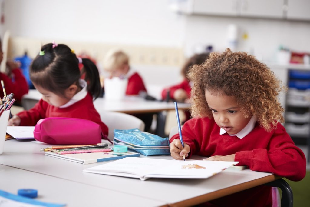 Young schoolgirl wearing school uniform sitting at a desk in an infant school classroom drawing, close up